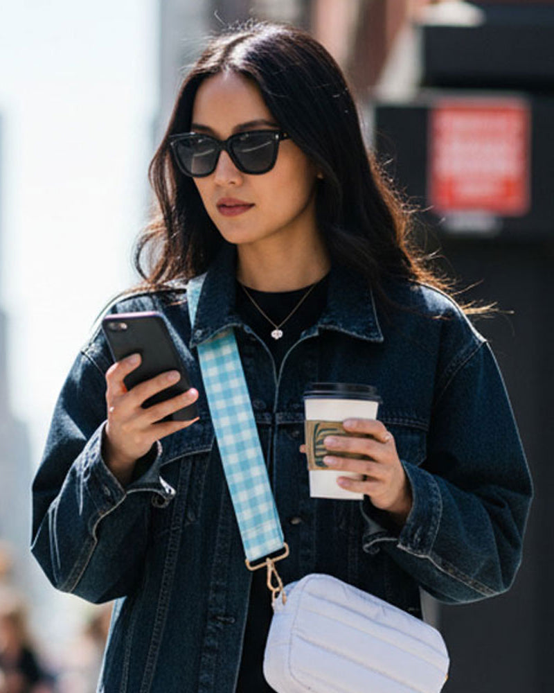 Woman in a denim jacket using a phone and holding a coffee cup on a city street. Wearing a white crossbody bag with a aqua plaid bag strap.