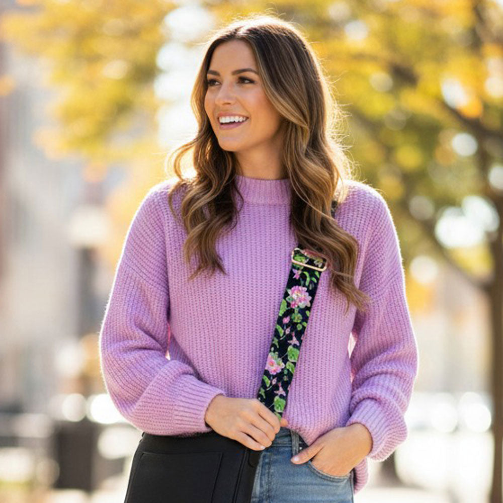 Woman wearing a purple sweater with a black ground floral strap with a black crossbody bag, standing outdoors with blurred trees in the background.