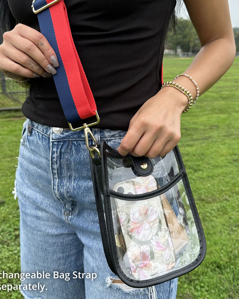 Person holding a clear bag with a red and blue strap, standing in a grassy field.