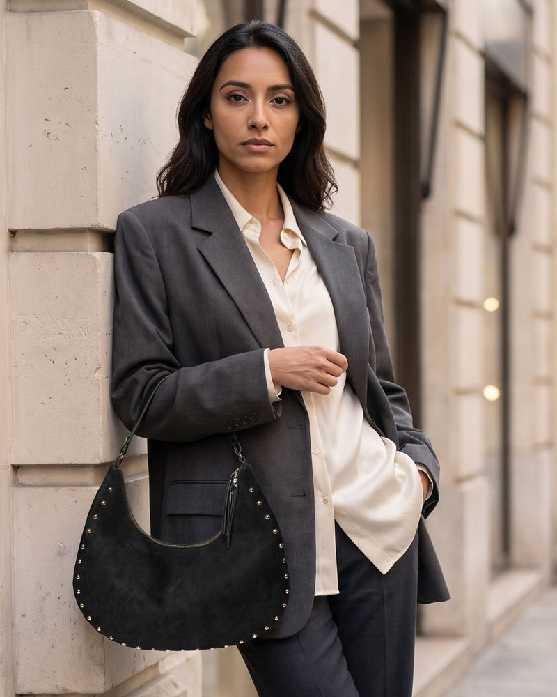 A woman stands on a city sidewalk against a beige stone building, wearing a dark gray blazer over a cream button-up shirt and dark trousers, while carrying a black crescent-shaped suede handbag with gold-tone studs.