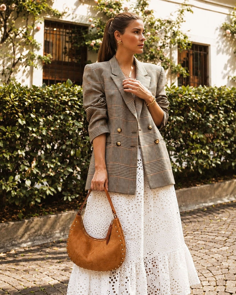 Woman in a plaid blazer and white dress holding a camel vegan suede with studs handbag on a street.