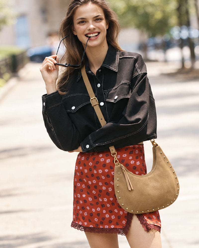 A woman stands on a sunlit city sidewalk wearing a black denim jacket, a red floral skirt, and carrying a tan suede crescent shoulder bag with silver studs and a tan strap.