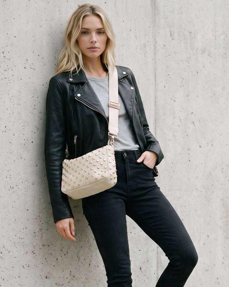 A blonde woman posing against a textured concrete wall, wearing a black leather moto jacket, black jeans, and a gray t-shirt, while carrying a cream vegan suede crossbody bag with a studded pattern and solid bag strap.