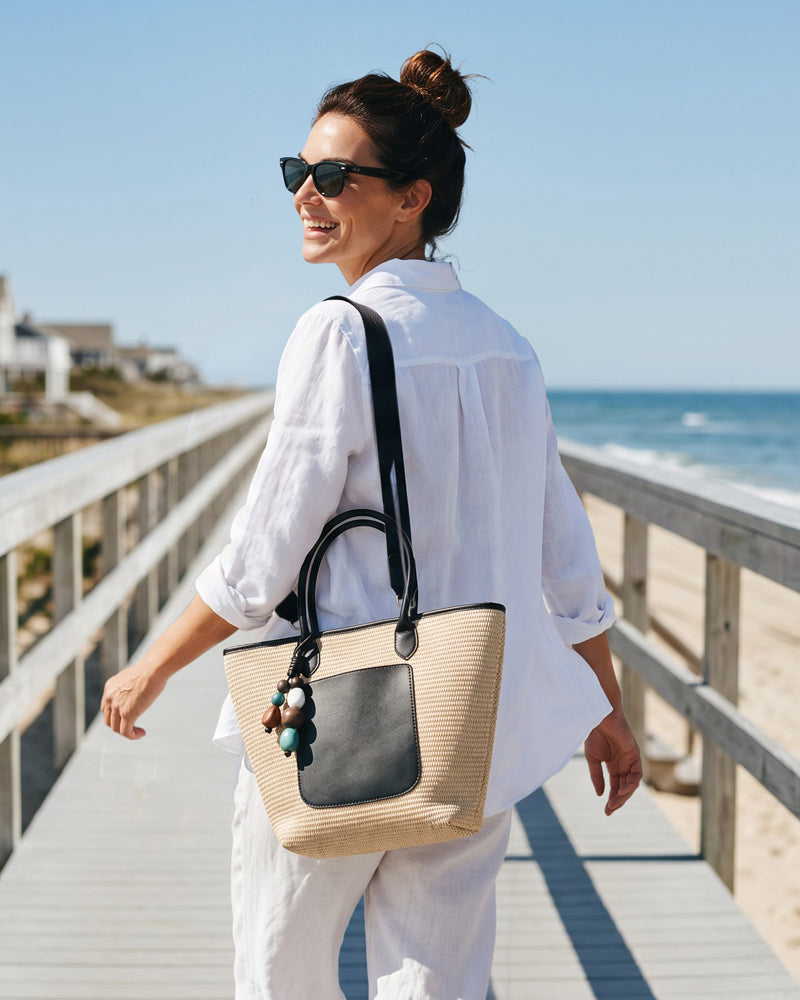 Woman walking on a beach boardwalk with a raffia tote, wearing sunglasses and a white outfit.