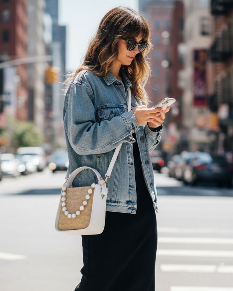 Woman in a denim jacket using a phone on a city street. Wearing a raffia handbag.