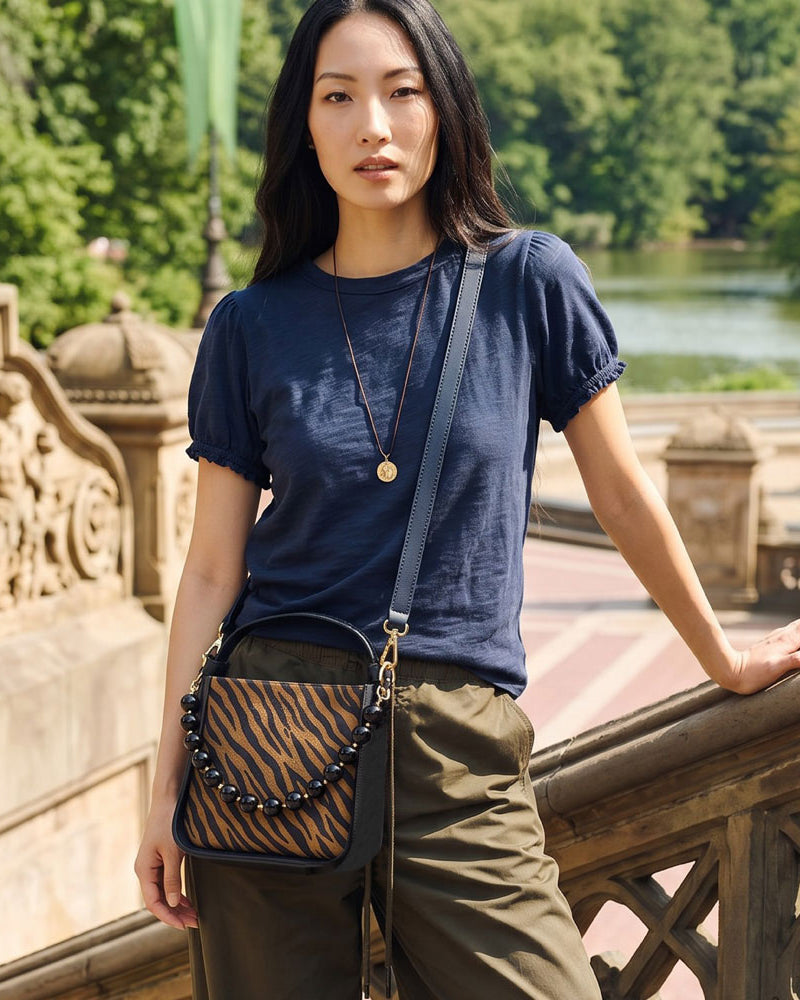 A woman stands on an outdoor stone bridge wearing a navy blue short-sleeve t-shirt, olive green pants, and a crossbody bag with a dark brown and black tiger-stripe pattern and a beaded strap.