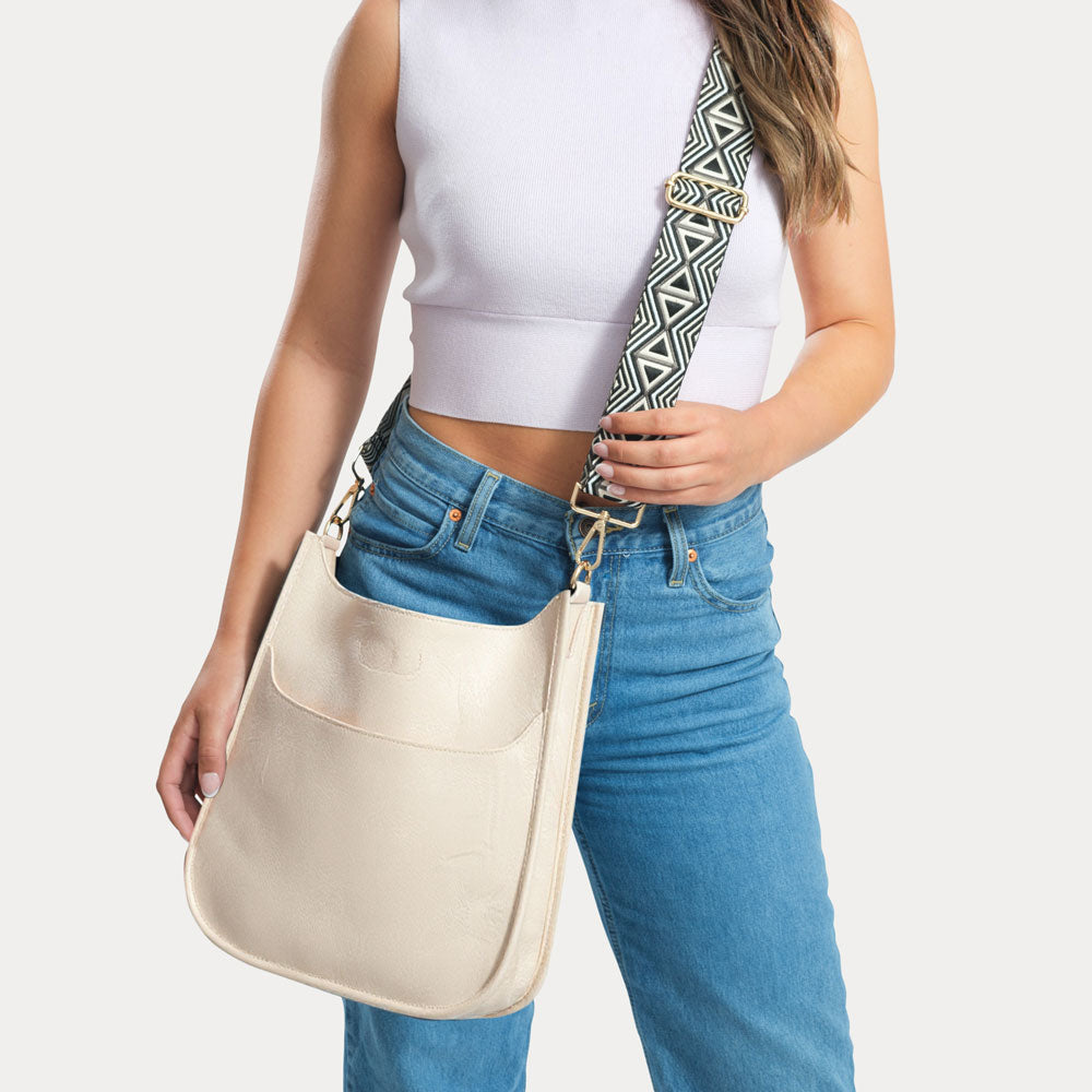 Woman wearing a white sleeveless top and blue jeans, holding a cream shoulder bag with a patterned strap.