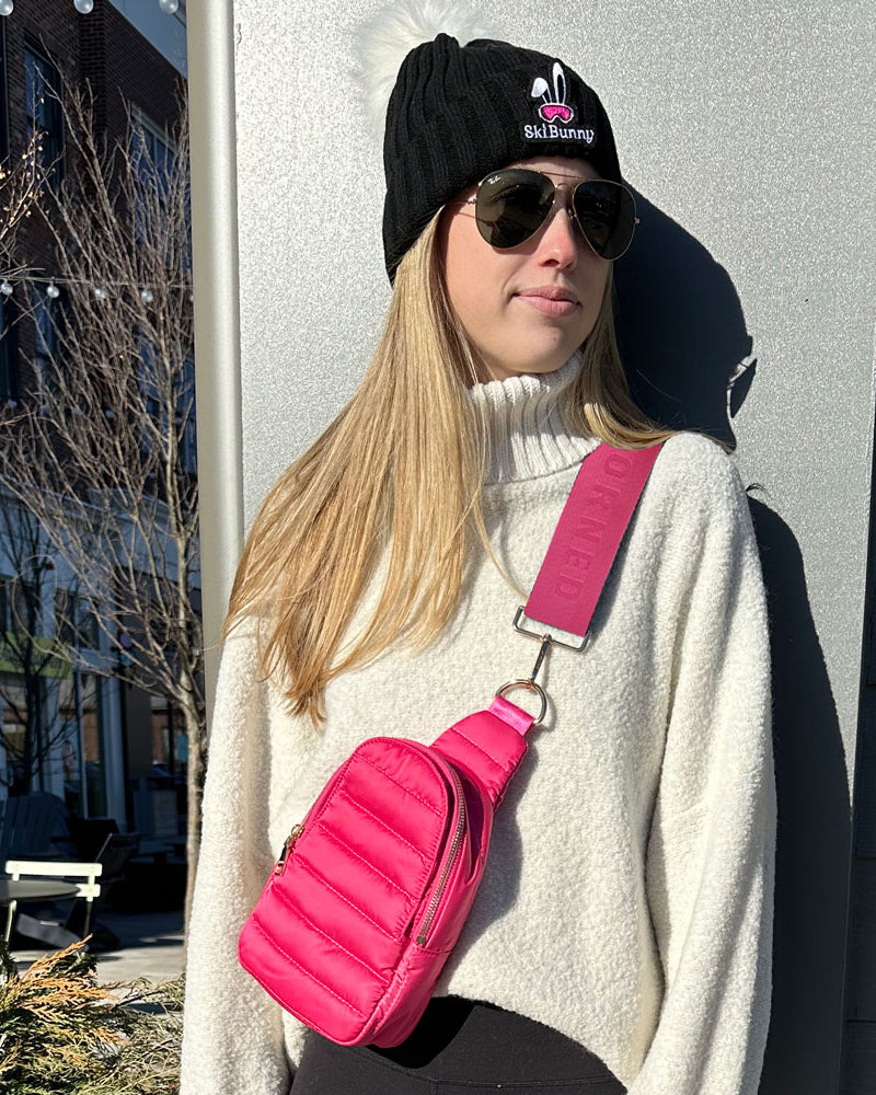 Person wearing a black beanie with a pink bunny design, sunglasses, and a white sweater with a pink crossbody bag.