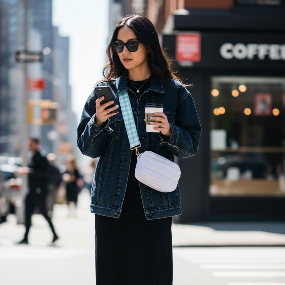 Woman in a city street holding a phone and a coffee cup, wearing a denim jacket and sunglasses. And wearing a white crossbody bag.