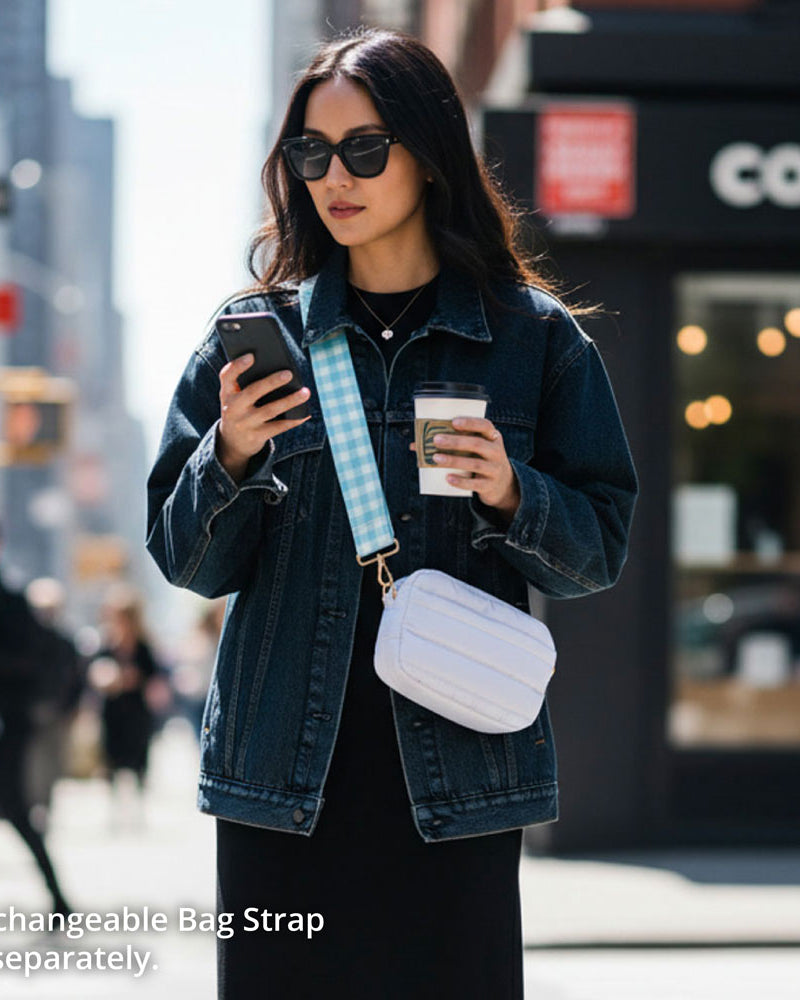 Woman in a city street holding a phone and a coffee cup, wearing a denim jacket and sunglasses. And wearing a white crossbody bag.