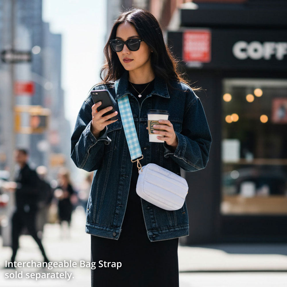Woman in a city street holding a phone and a coffee cup, wearing a denim jacket and sunglasses. And wearing a white crossbody bag.