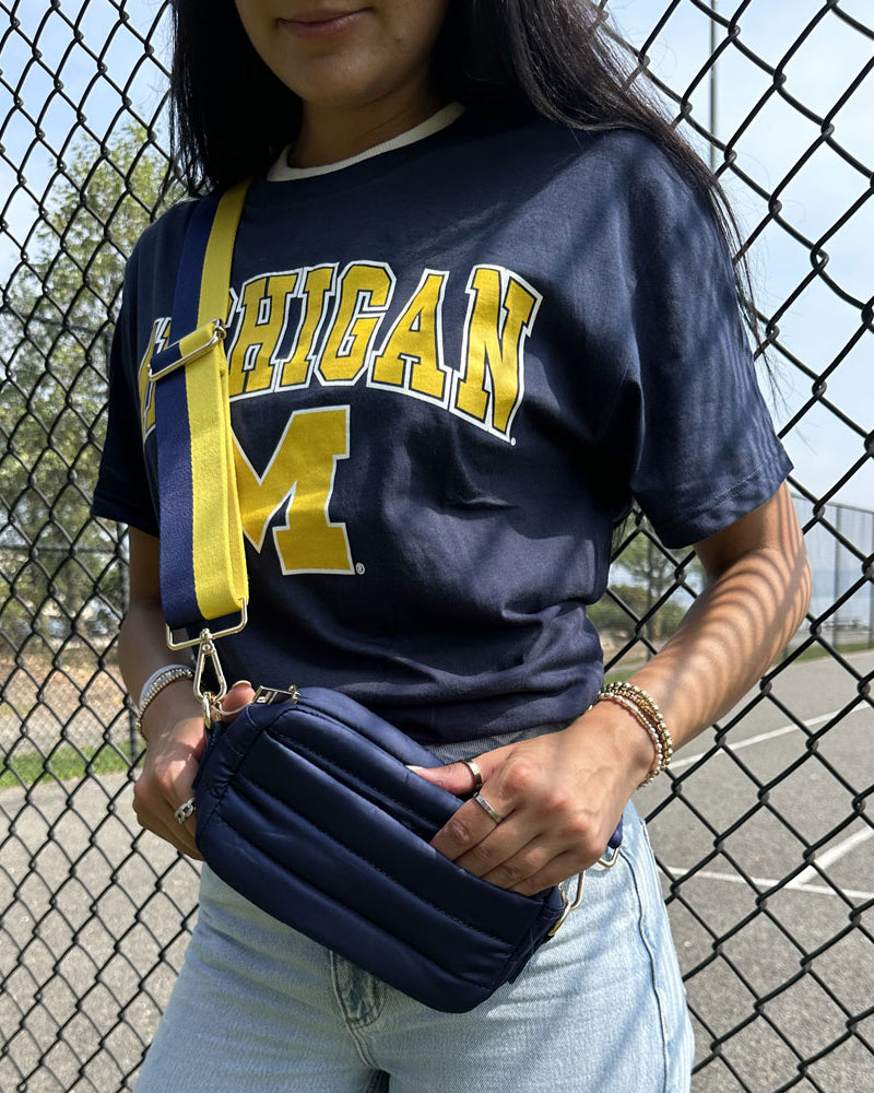 woman wearing maize and azure striped strap and Ella Quilted Puffer bag in Navy, standing in front of a chain link fence
