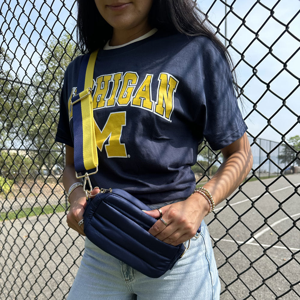 woman wearing maize and azure striped strap and Ella Quilted Puffer bag in Navy, standing in front of a chain link fence