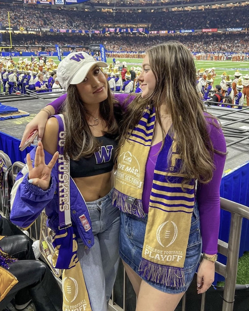2 woman posing in a football stadium. One is wearing the white and purple game day striped bag strap