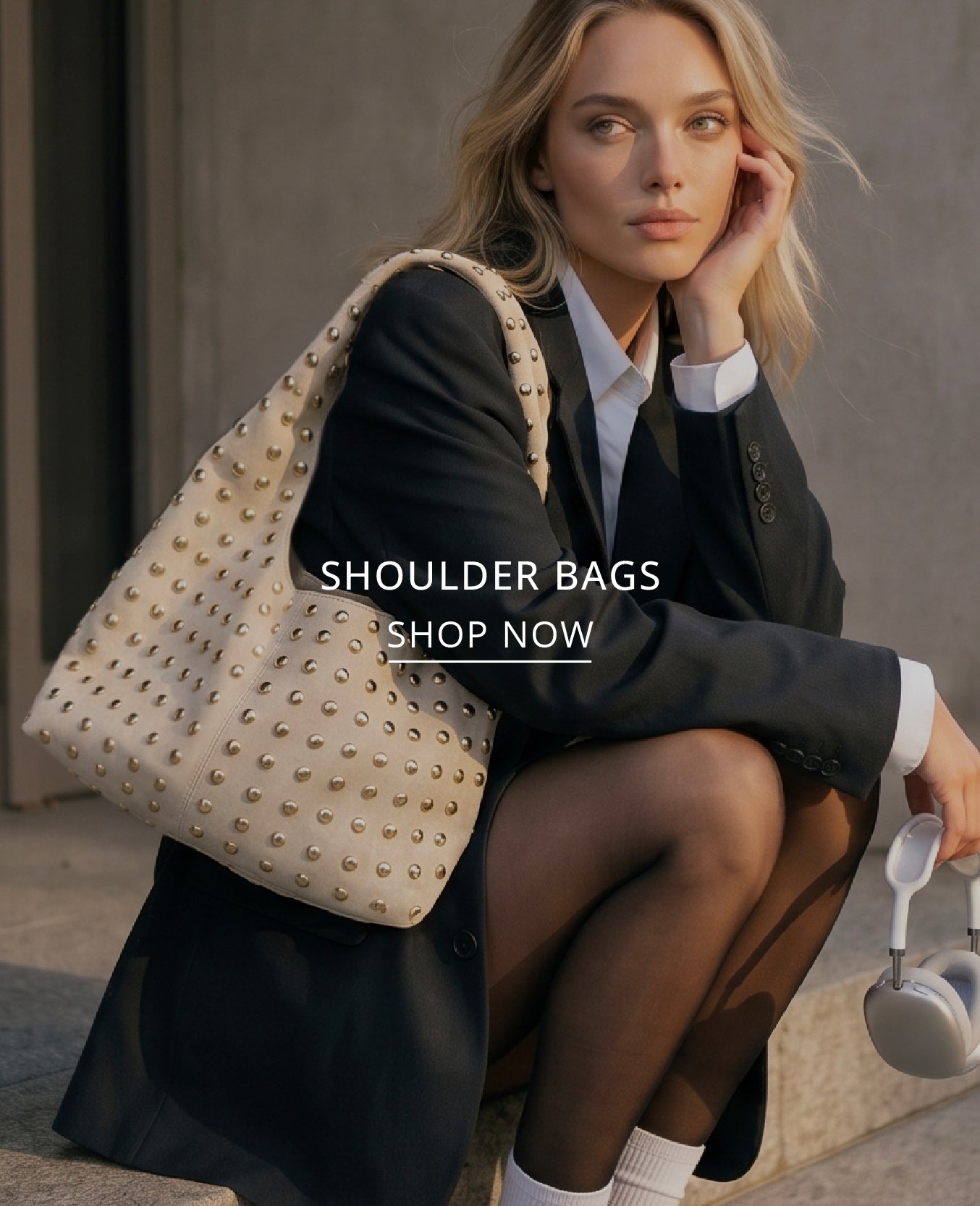 A woman in a black blazer and white shirt sits on stone steps, wearing a large cream shoulder bag covered in gold studs. The text 'SHOULDER BAGS SHOP NOW' is overlaid on the center of the image.