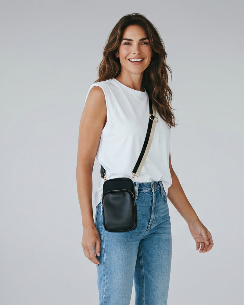 Woman wearing a white sleeveless top and blue jeans with a black crossbody bag on a plain background