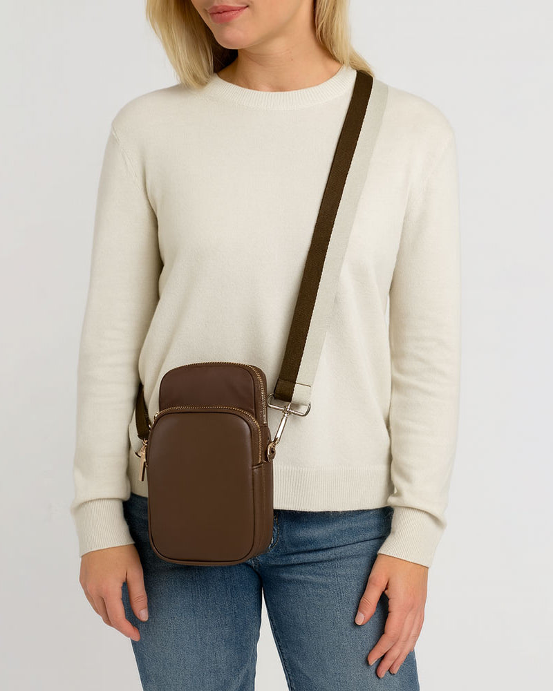 Person wearing a beige sweater and blue jeans with the Chocolate Janet crossbody bag on a light gray background