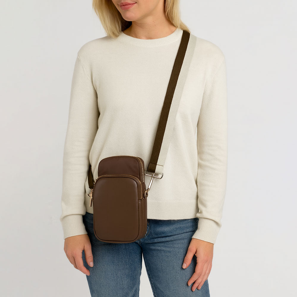 Person wearing a beige sweater and blue jeans with the Chocolate Janet crossbody bag on a light gray background