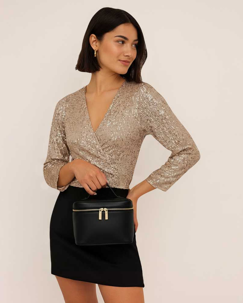 Woman wearing a sparkly top and black skirt holding a black bag against a plain background