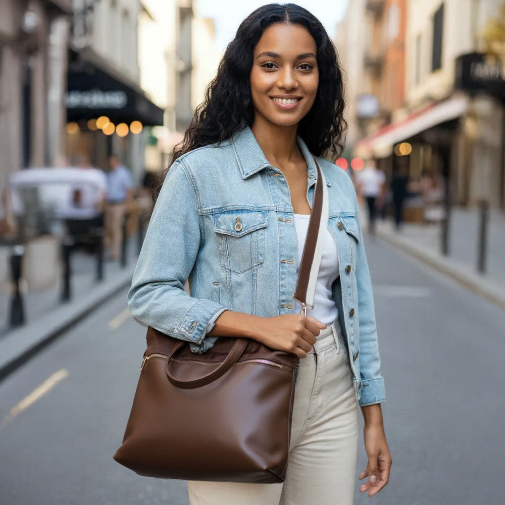 Woman in a denim jacket and white pants holding a brown leather bag on a city street.