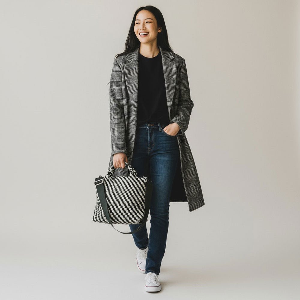 Woman holding a army and ecru woven neoprene tote, wearing a long coat and jeans, on a plain background