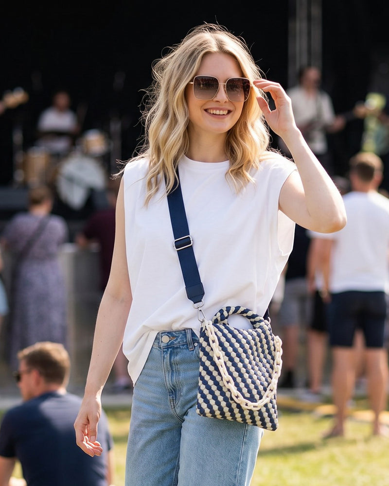 Woman in a white sleeveless top and blue jeans with a striped bag, standing outdoors with blurred people in the background.