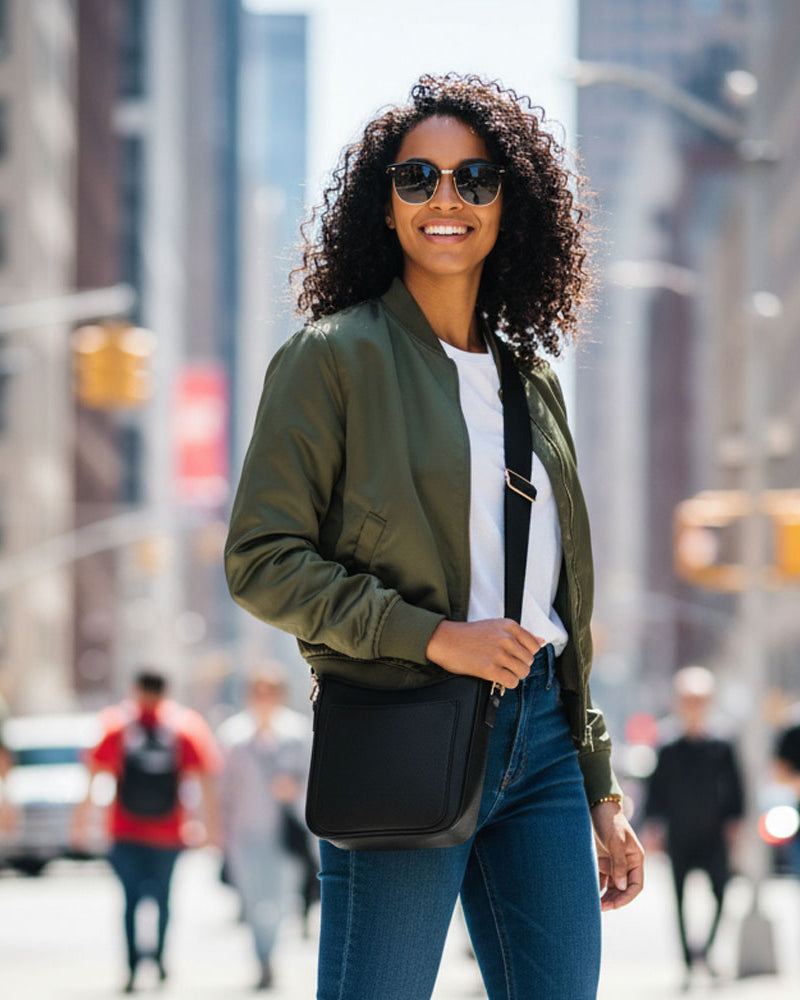 Woman walking on a city street wearing sunglasses and a green jacket. She is wearing a black crossbody bag.
