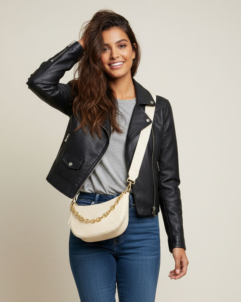 Woman wearing a black moto jacket, grey tee, denim, and a cream crossbody bag with chain detail.