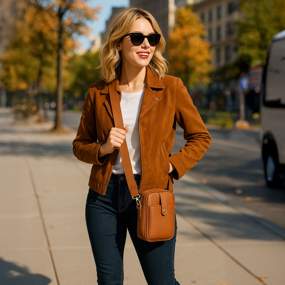 Woman in a brown jacket and sunglasses walking on a sidewalk with trees and buildings in the background wearing a camel crossbody bag