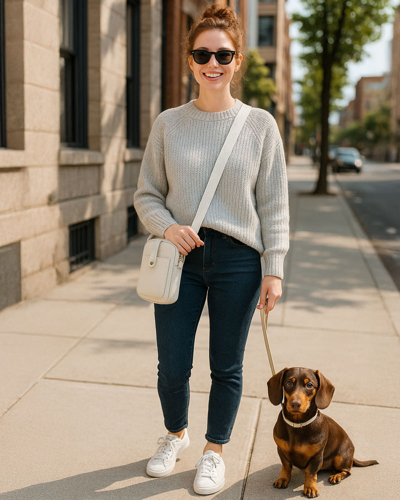 Woman walking a small dog on a city street. Wearing a cream crossbody bag.