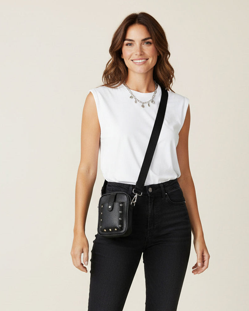 Woman wearing a white sleeveless top and black pants with a black crossbody with silver studs bag on a beige background