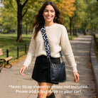 Woman walking outdoors in a park, wearing a cream sweater and black skirt with a black handbag.  Text: Note: Strap shown in photo not included.  Please add a bag strap to your cart.