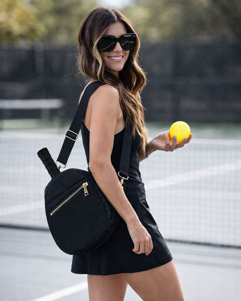 Woman holding a yellow tennis ball on a tennis court with a black pickleball paddle bag.