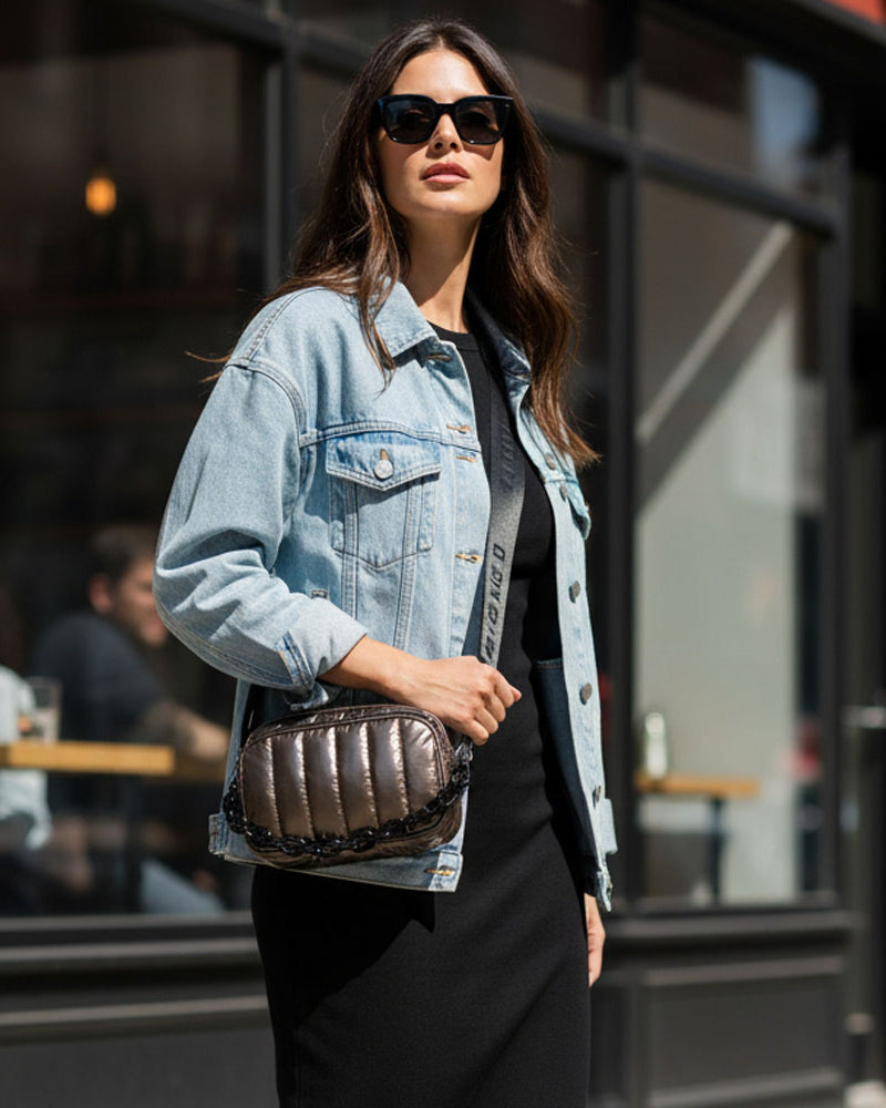 Woman wearing a denim jacket and sunglasses, holding a bronze metallic handbag, standing in an urban setting.