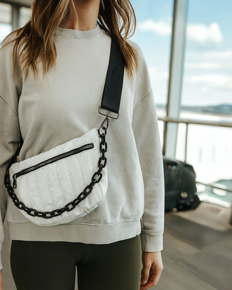 Person wearing a light gray sweatshirt with a white bag featuring black chain details, standing in an airport.