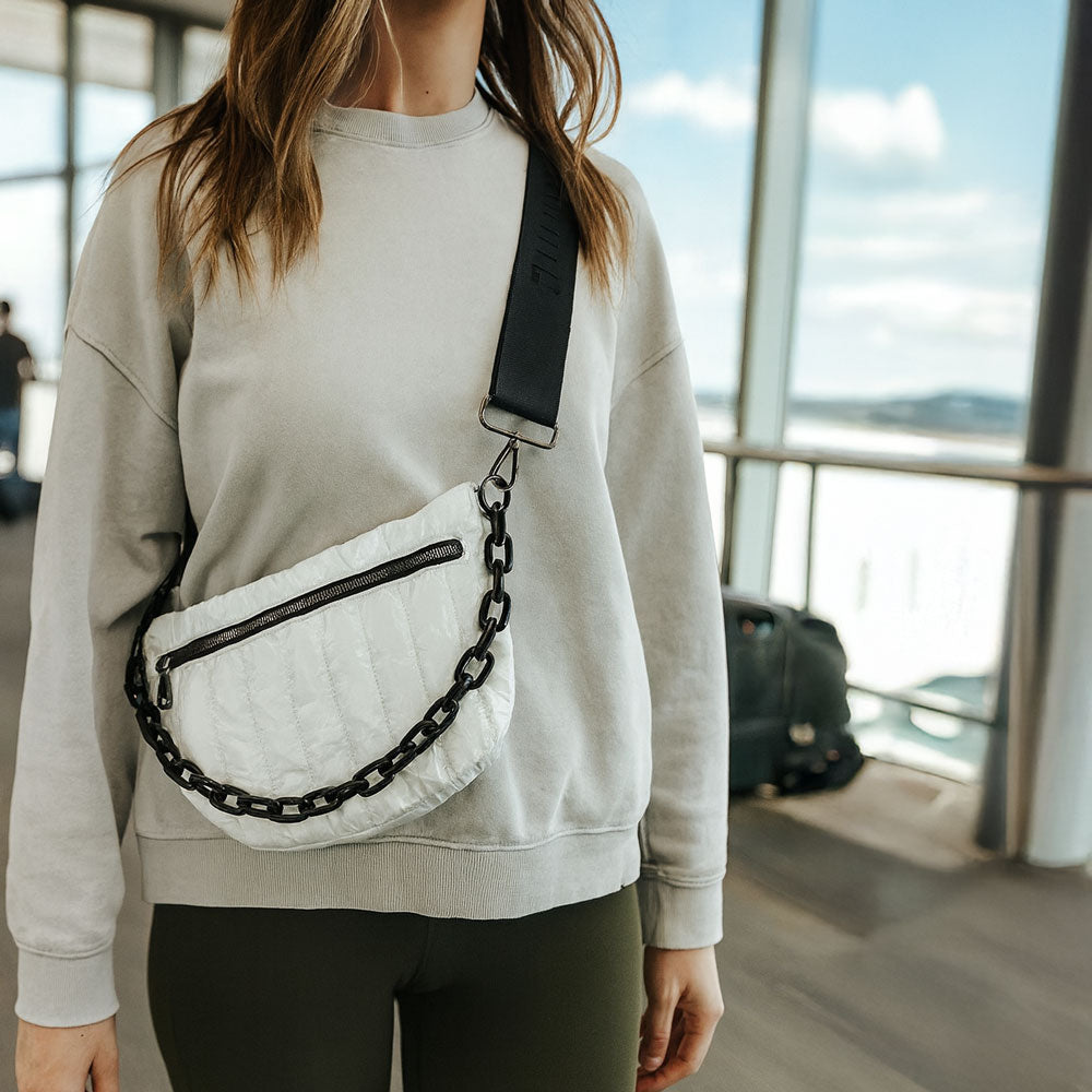 Person wearing a light gray sweatshirt with a white bag featuring black chain details, standing in an airport.