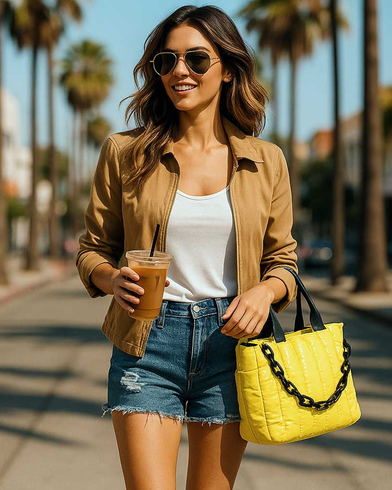 Woman walking on a sunny street holding a yellow tote and a drink.