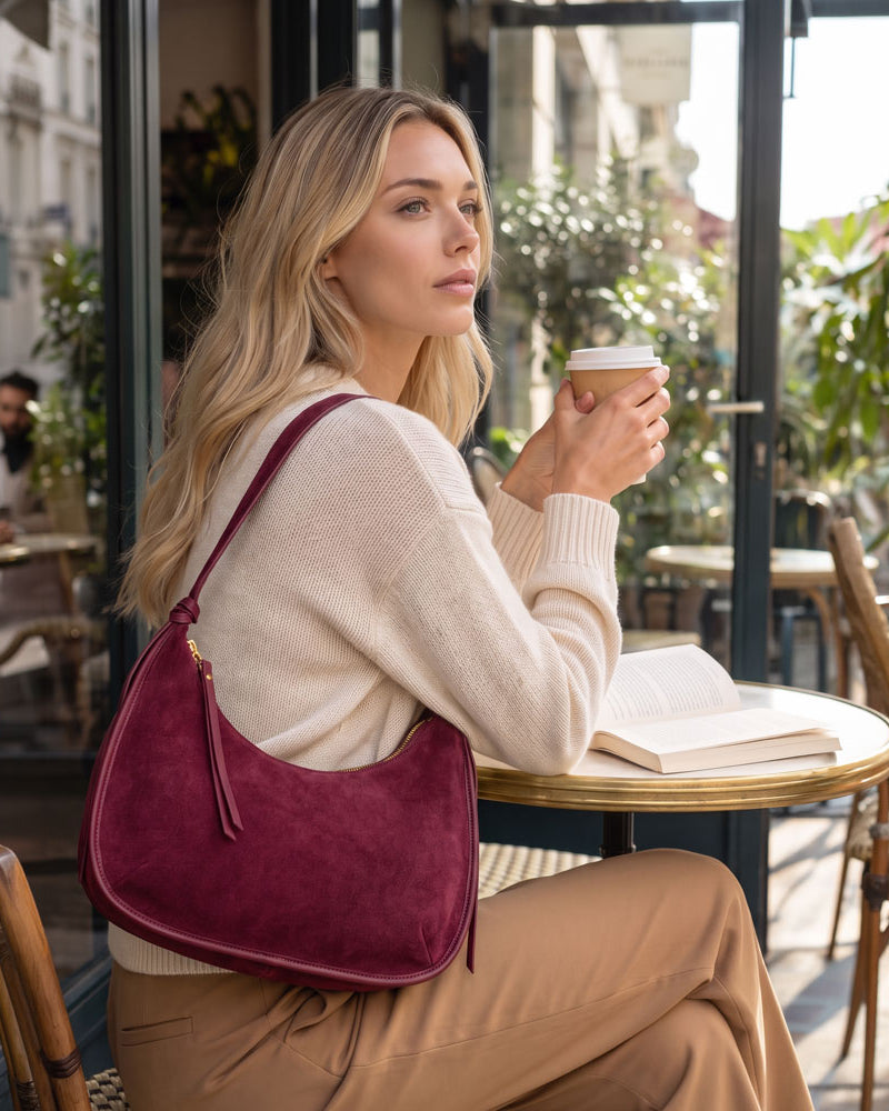 A woman sitting at a cafe table holding a coffee cup, wearing a cream knit sweater, tan pants, and carrying a burgundy suede shoulder bag.