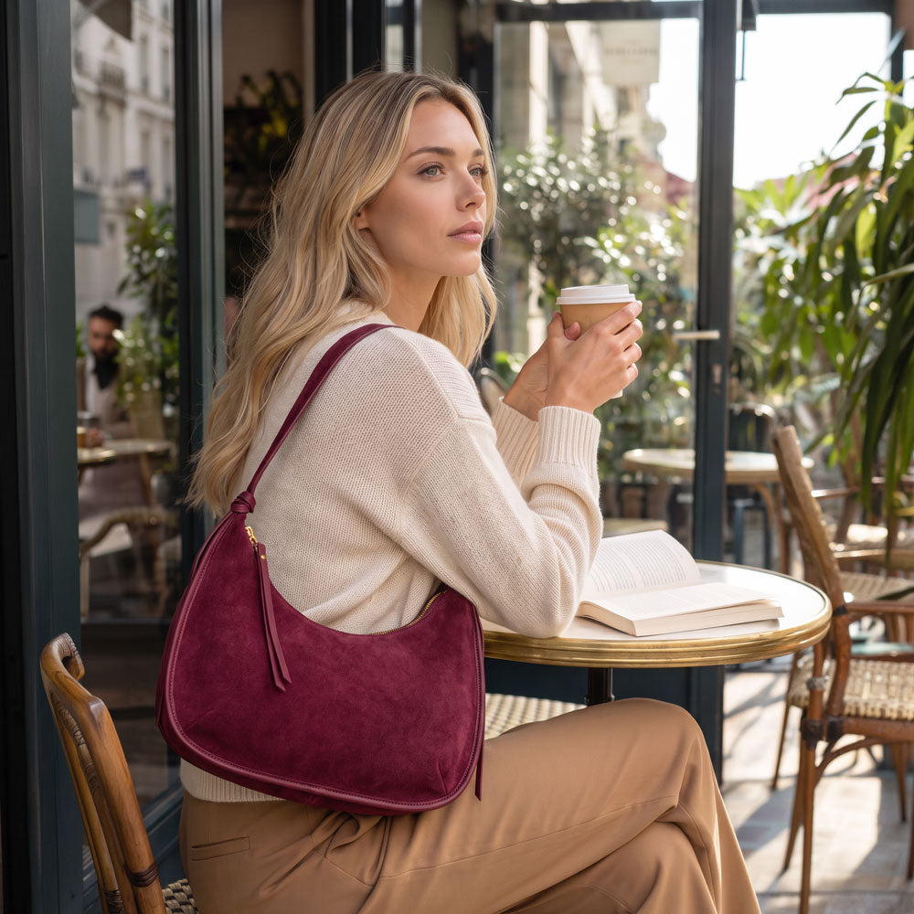 A woman sitting at a cafe table holding a coffee cup, wearing a cream knit sweater, tan pants, and carrying a burgundy suede shoulder bag.