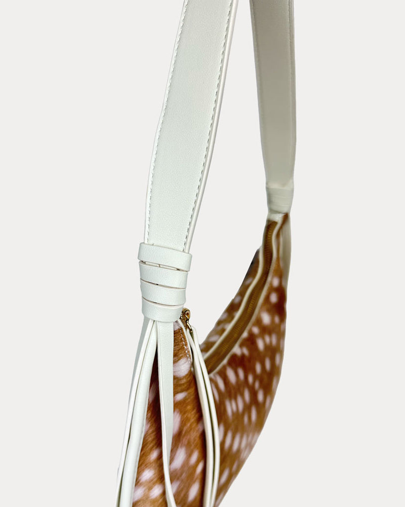 A close-up of a shoulder bag featuring a brown and white deer animal print body with a cream vegan leather strap and tassel detail, set against a solid light gray background.
