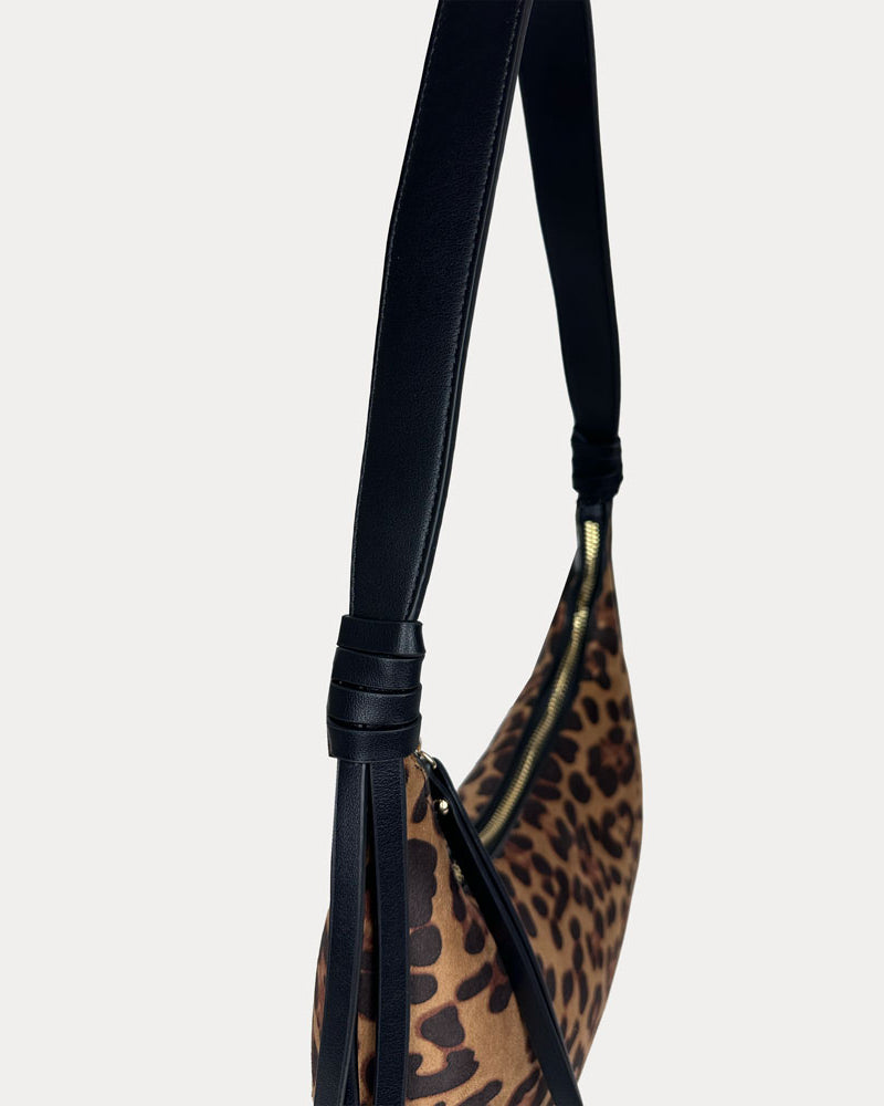 A close-up of a shoulder bag featuring a brown and black leopard print pattern with a black vegan leather strap set against a solid light gray background.