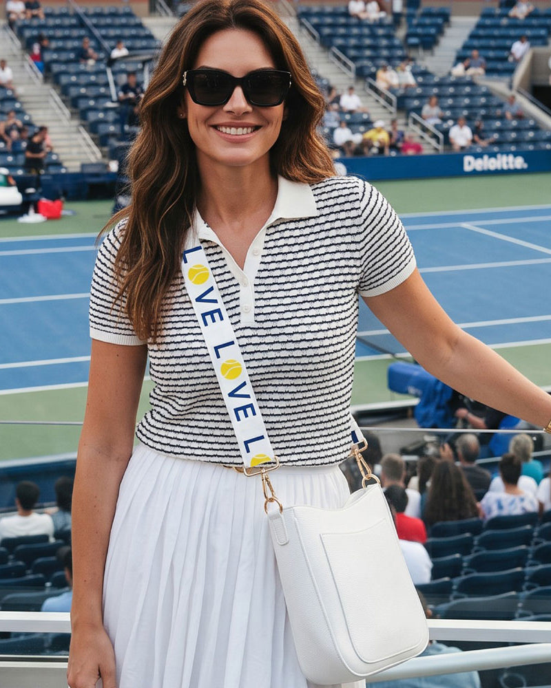 A woman stands in the standard a tennis match wearing a navy and white striped short-sleeve polo shirt, a white pleated skirt, black sunglasses, and a white crossbody bag with white 'LOVE' strap. The background shows stadium seating and a blue tennis court.