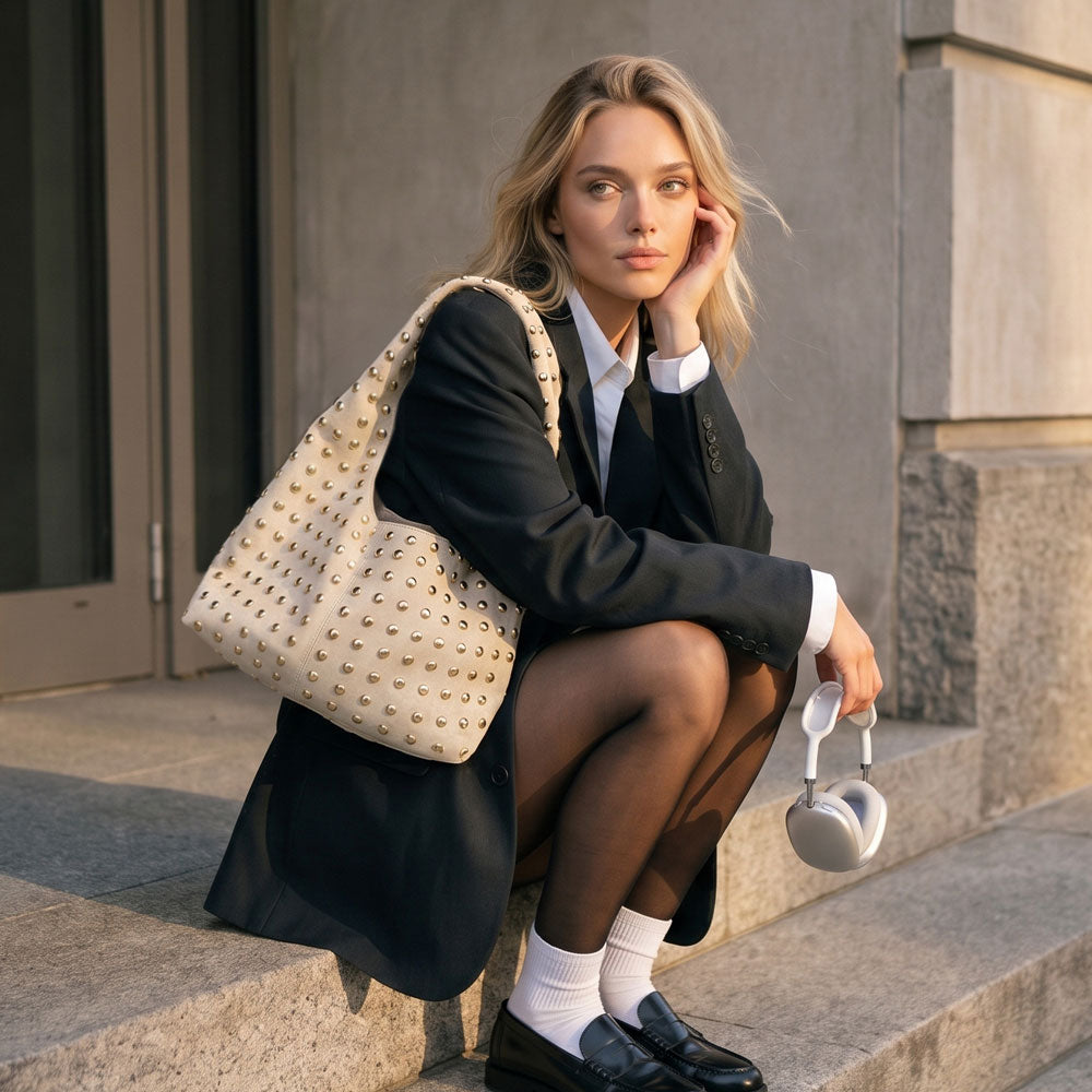 Woman sitting on stone stairs on the city. Wearing a grey overcoat, white button down shirt, black skirt, black tights, white socks with black loafers.  She is wearing a cream studded hobo bag