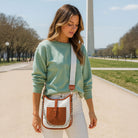 Woman in a green sweater and white pants holding a canvas and camel handbag outdoors.