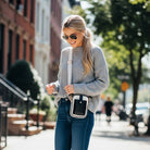 Woman walking on a city street wearing sunglasses and a gray sweater with neutral canvas with black vegan leather details handbag.