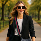 Woman wearing sunglasses and a black blazer with a green, red, and black striped bag strap with a black handbag in a park setting