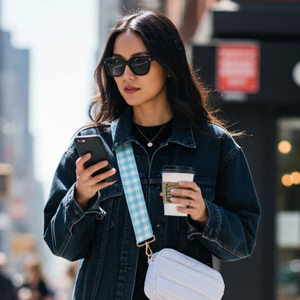 Woman in a denim jacket using a phone and holding a coffee cup on a city street. Wearing a white crossbody bag with a aqua plaid bag strap.