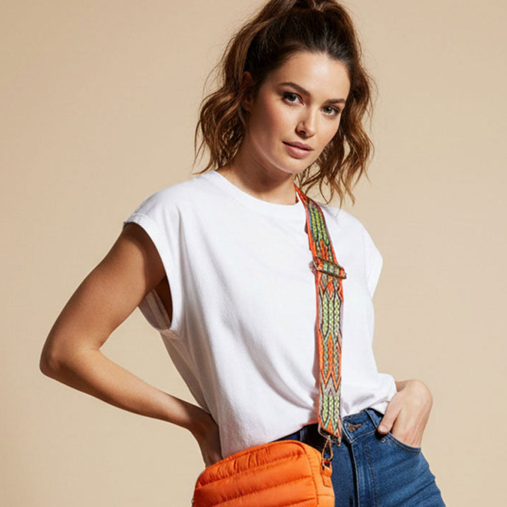 Woman wearing a white t-shirt and blue jeans with an colorful bag strap with an orange on a beige background