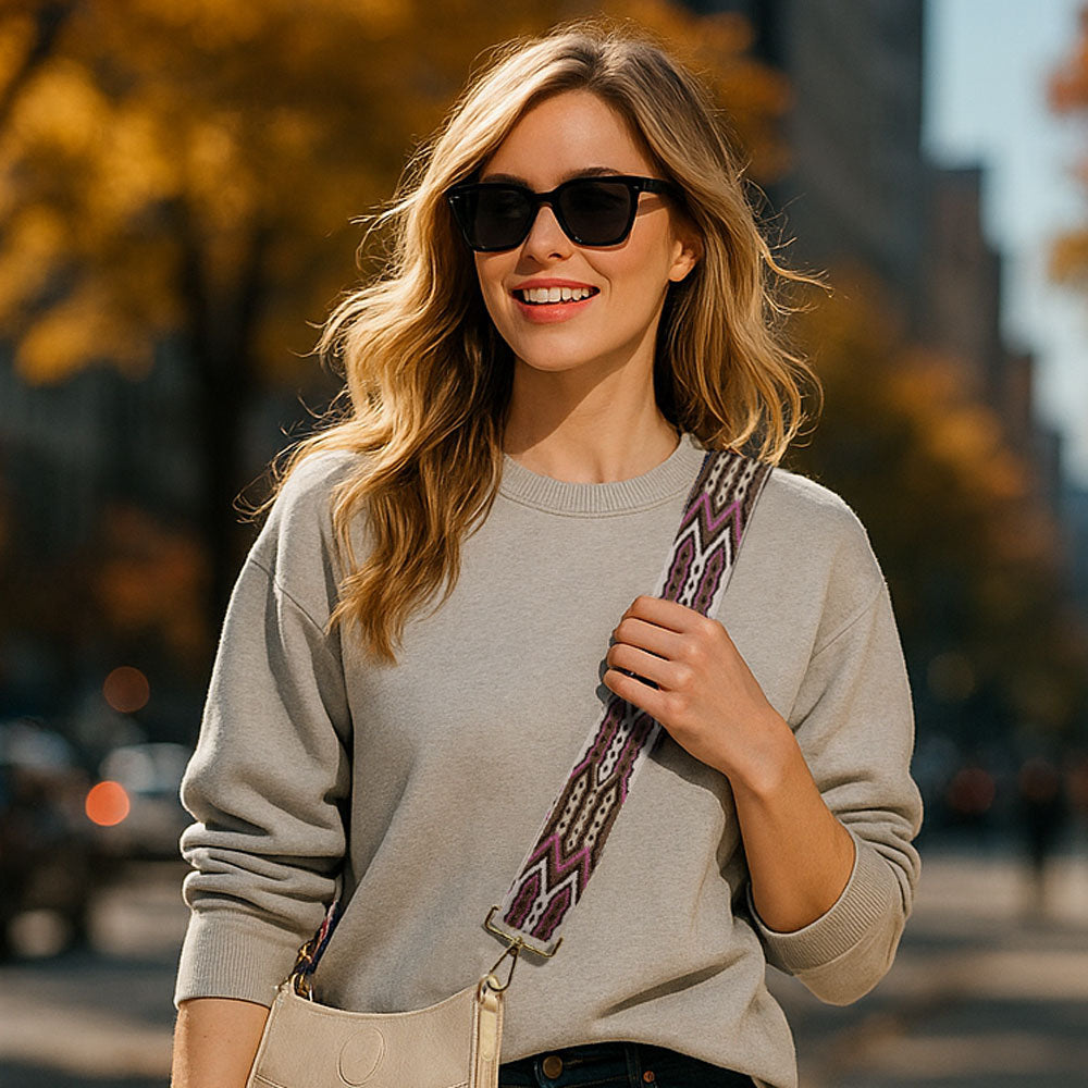 Woman wearing sunglasses and a gray sweater with a colorful strap, standing outdoors.
