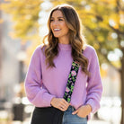 Woman wearing a purple sweater with a black ground floral strap with a black crossbody bag, standing outdoors with blurred trees in the background.